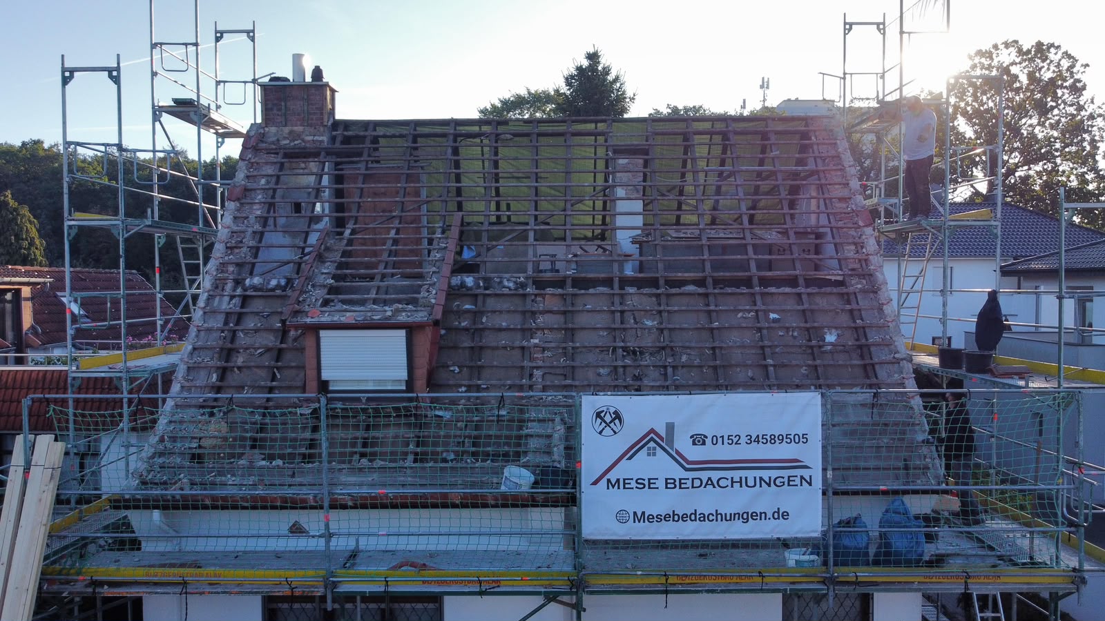 Dachsanierung an einem Wohnhaus: freigelegte Holzlattung, Gerüst mit Grün und Baustellenschild von Mese Bedachungen