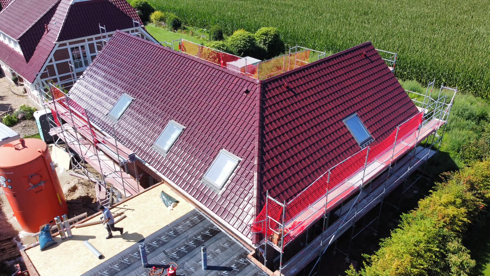 Luftaufnahme eines fertig gedeckten Dachs mit glänzenden dunkelroten Dachziegeln, vier Dachflächenfenstern und abgedichtetem Flachdachanbau